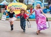 Three children in costumes walk together holding trick-or-treat bags at SeaWorld's Halloween event, with colorful decorations in the background.