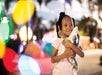A young girl stands outdoors at dusk, smiling and hugging a stuffed toy dolphin, with colorful blurred lights in the background.