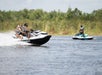 Two people ride a jet ski across a body of water while another person rides a separate jet ski in the background, with reeds and trees visible on the shore.
