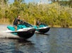 Two people ride teal and black jet skis side by side on a lake with trees and shrubs in the background.