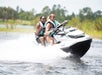 Two people wearing life jackets ride a white and black jet ski on the water, creating a splash, with greenery visible in the background.