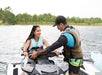 A woman on a jet ski smiles at a man who appears to be giving her instructions. Both are wearing life jackets near a river or lake with trees in the background.