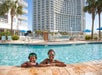 Two children wearing swim goggles smile while resting on the edge of a swimming pool, with a tall hotel building and palm trees in the background.