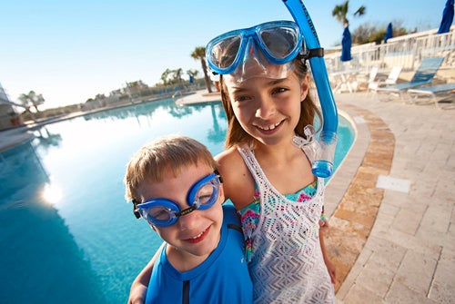 Two children wearing swim goggles and snorkels stand by a pool on a sunny day, smiling at the camera.