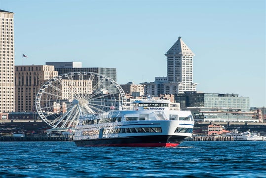 A white Argosy cruise ship on the water with a city skyline, Ferris wheel, and tall buildings in the background under a clear blue sky.