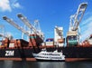A white yacht passes in front of a large cargo ship loaded with shipping containers, with cranes positioned overhead against a blue sky.