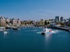 A passenger ferry travels across a harbor with a cityscape, buildings, and marina visible in the background under a clear blue sky.