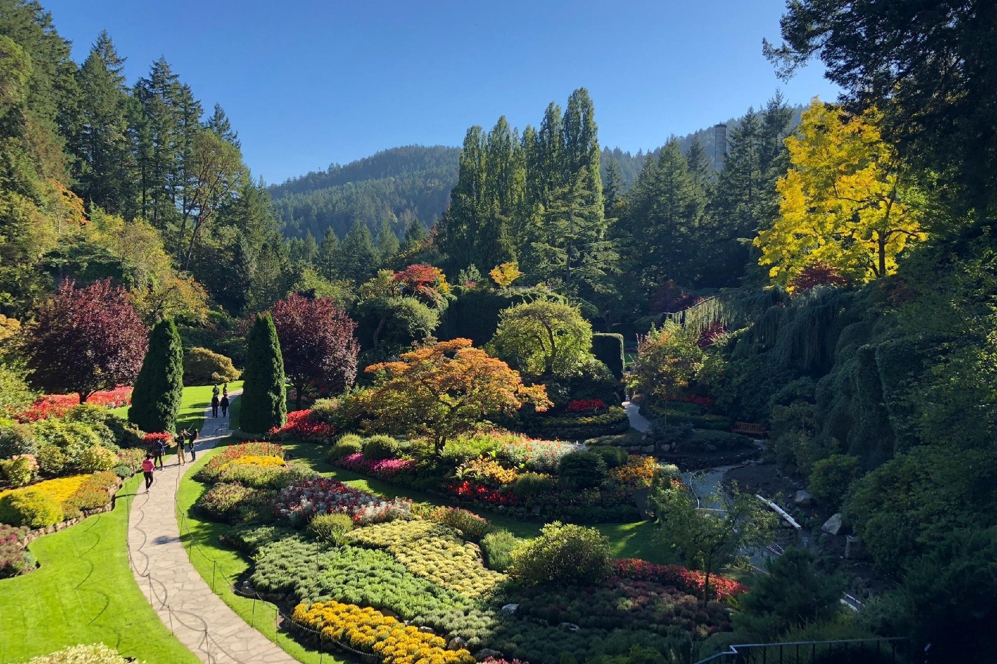 A landscaped garden with colorful flower beds, green lawns, winding paths, and tall trees under a clear blue sky, surrounded by forested hills.