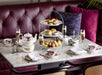 A set table for afternoon tea with floral china, a teapot, sandwiches, scones, pastries, and glasses of champagne on a marble surface with a burgundy sofa in the background.