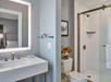 Modern bathroom with a white sink and illuminated mirror, glass shower, and toilet. A small shelf holds toiletries, a picture hangs on the wall, and a vase with flowers is near the toilet.