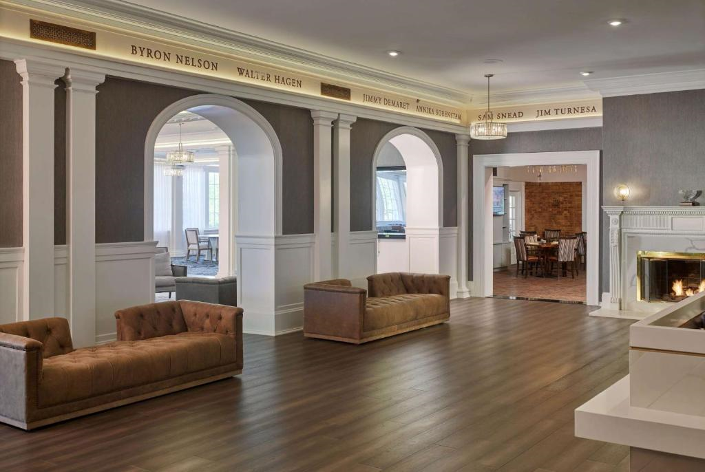 A spacious lobby with wooden flooring, brown sofas, and a fireplace. Arched openings lead to another room. Names are displayed on the upper wall.