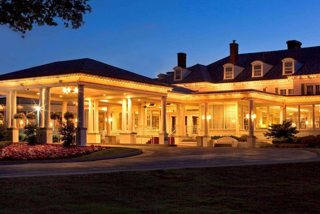 A grand hotel entrance illuminated at dusk with warm lights, surrounded by pillars, flowers, and trees.