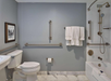 An accessible bathroom with grab bars, a toilet, a towel rack with white towels, a bathtub with a showerhead, and a framed picture on the wall.