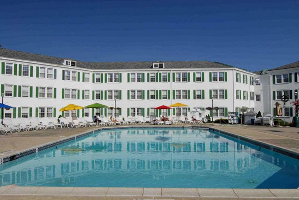 A large, rectangular outdoor pool surrounded by white lounge chairs and green-trimmed windows on a white building, under a clear blue sky. Colorful umbrellas provide shade.