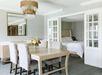 A dining area with a light wood table and six upholstered chairs. There's a chandelier above the table and a bouquet on top. Glass doors open to a bedroom with a white bed.
