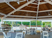 Outdoor pavilion with wooden ceiling, tables covered in striped cloths, a decorative boat on the left, and neatly trimmed hedges in the background.