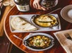 A table with a bottle of wine, two iron skillets containing melted cheese dishes, and a stack of tortillas on a plate. A hand holds a glass of red wine.