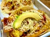 Close-up of tacos with scrambled eggs, shredded cheese, tortilla chips, and avocado slices on a tray.
