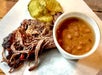 Pulled pork with pickle slices on a plate, accompanied by a cup of baked beans.