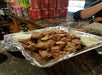 A tray of grilled sausages cut into pieces, served with dipping sauces, sits on a counter lined with aluminum foil. Stacks of canned drinks are in the background.