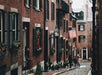 A narrow cobblestone street between red brick buildings, with black shutters and balconies. An American flag hangs from one building. Two people walk along the sidewalk.