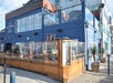 A blue building with a restaurant featuring outdoor seating enclosed by wooden and glass panels. People sit at tables. A staircase leads to a second-floor patio with string lights.