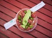 A poke bowl with sliced tuna, avocado, green onions, and sesame seeds on a red wooden table, with chopsticks beside it.