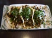 Three tacos on a plate topped with chopped cilantro and onions, featuring various fillings on a decorative paper-lined surface.