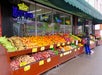 A sidewalk fruit and vegetable stand displays bananas, oranges, sweet potatoes, and more. A person with a backpack browses the selection under a green awning.
