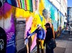 Two people are examining a colorful mural on an urban street. One person is pointing at text on the mural.