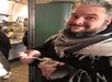 Person in a scarf eating food from a small cup with a fork, standing indoors near a food counter.