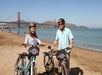 Two people with bicycles stand on a sandy beach near the water, with the Golden Gate Bridge and hills visible in the background under a clear sky.