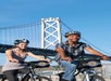 Two people wearing helmets ride bicycles in front of a large suspension bridge on a clear, sunny day.