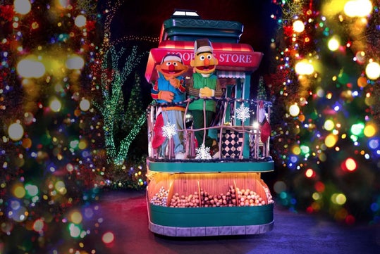 Two Sesame Street characters stand on a festive holiday float decorated with lights, ornaments, and snowflakes, surrounded by colorful, blurred Christmas lights.