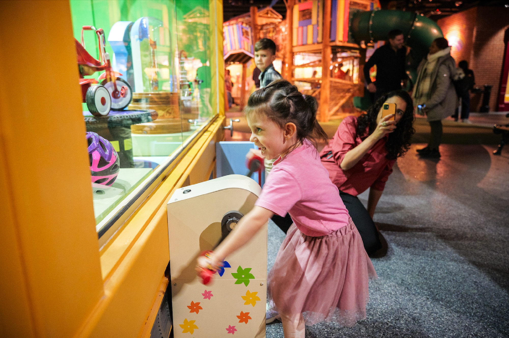 A young girl enthusiastically presses a button on an interactive display while a woman takes a photo. Other children and adults observe in a colorful, indoor playground.