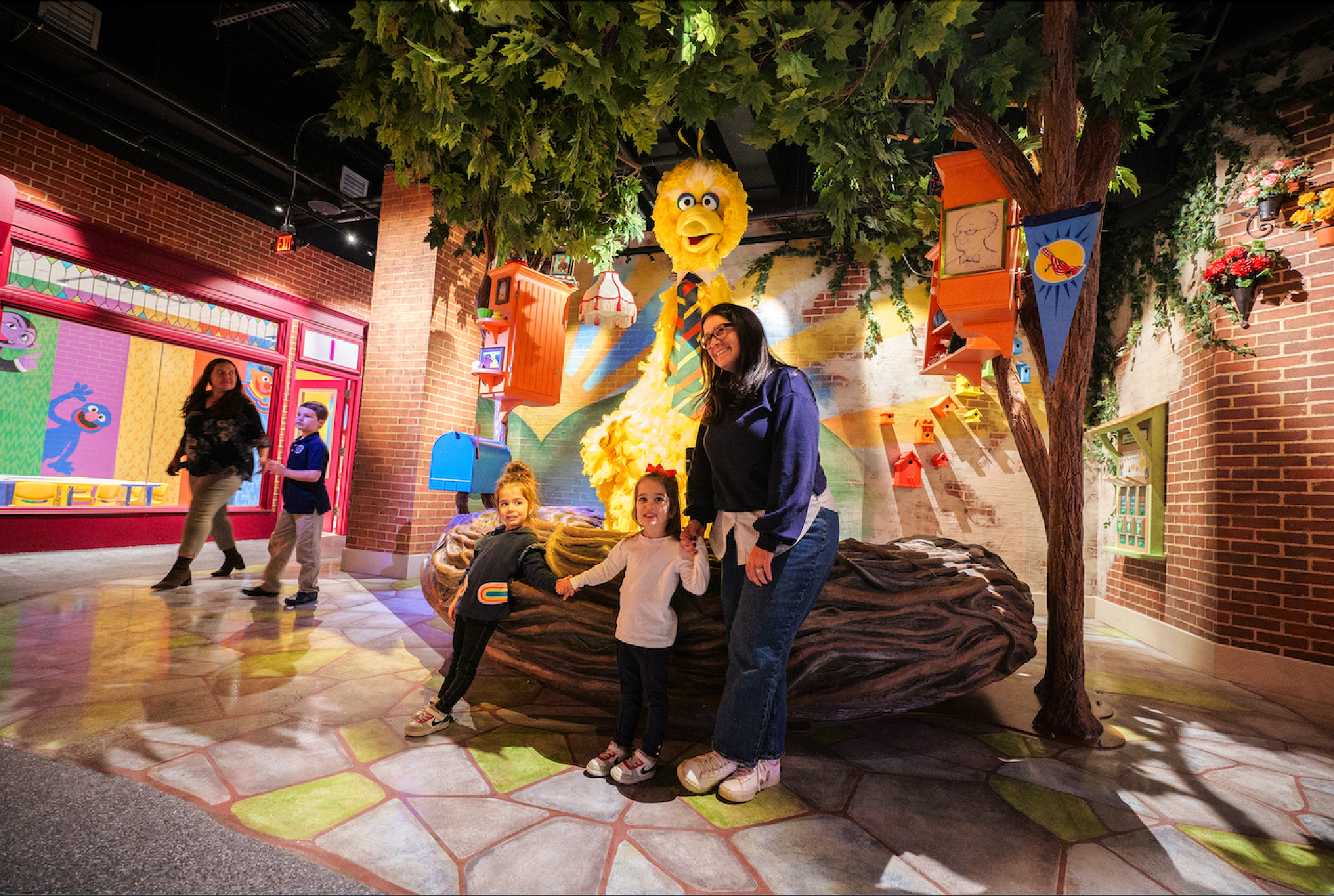 A woman and two children stand in front of a large yellow bird statue in a colorful, whimsical indoor setting with trees and decorations.