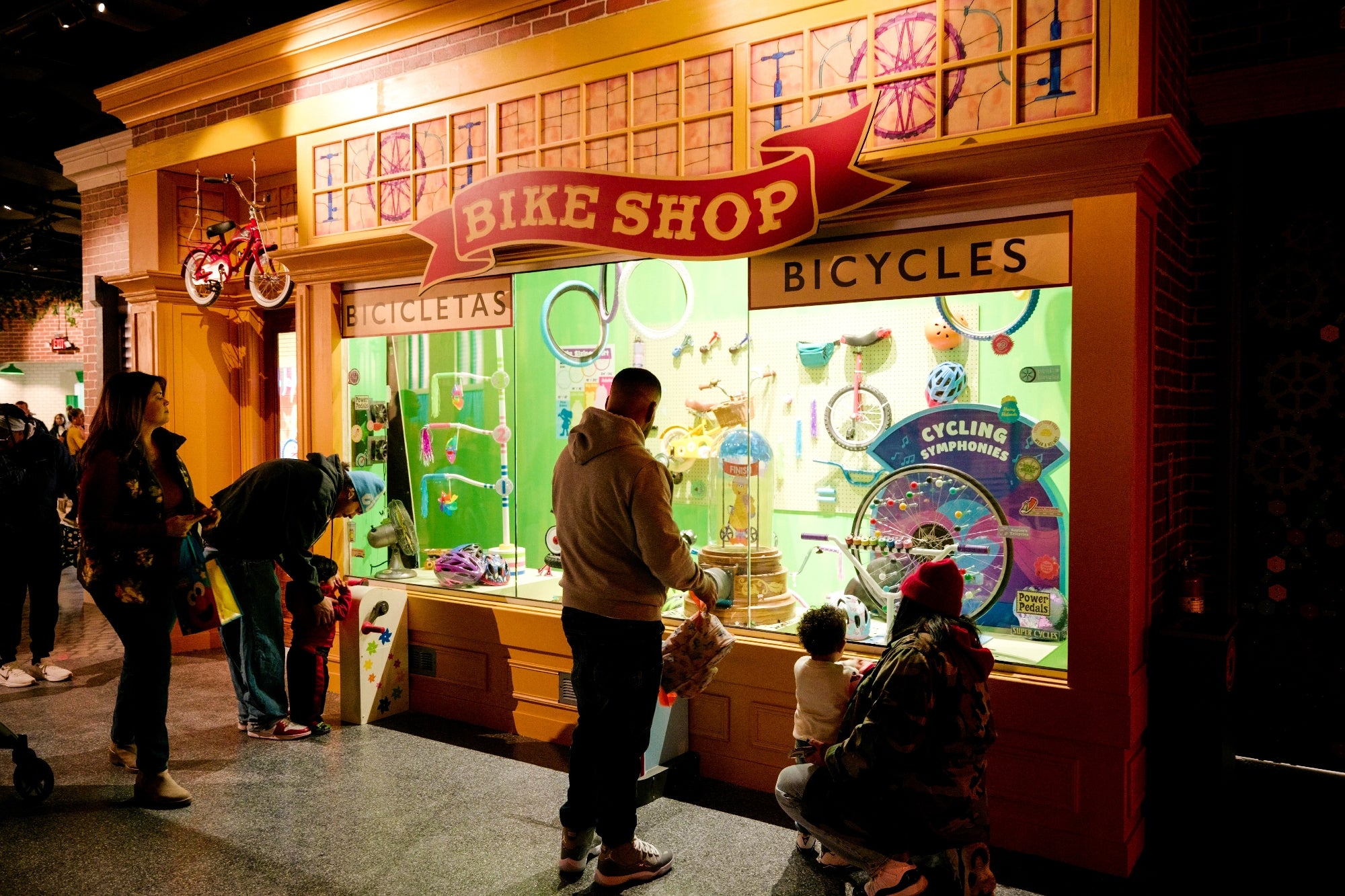 People visit a bike shop exhibit with bicycles and accessories displayed in a vibrant window under bright lights.