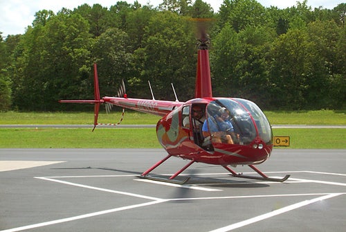 A red helicopter with two people inside is on the ground at a helipad, with grass and trees in the background.