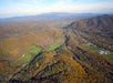 Aerial view of a rural landscape with rolling hills, forested areas, scattered farms, and a distant mountain range under a clear sky.