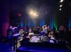 A musician performs on a piano under stage lights while a lively audience stands and watches in a dimly lit venue.