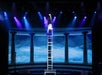 A performer does a handstand on top of a tall ladder on a stage with blue lighting and a backdrop of columns and clouds.