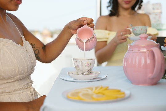Two women sit at a table having tea; one pours milk from a pink creamer into a teacup. Slices of lemon and a pink teapot are also on the table.