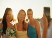 Three women pose together and smile for a photo outdoors at sunset, with water and sunlight in the background.