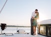A man and woman stand close together on a boat at sunset, with water and a distant shoreline in the background.