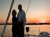 An older couple stands on a sailboat at sunset, looking at each other and smiling, with calm water and an orange sky in the background.
