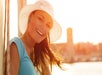 Woman wearing a white sunhat and blue shirt smiling outdoors with sunlit buildings in the background.