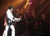 Performer in a white jumpsuit plays guitar on stage, with backup singers and musicians under colorful stage lights.