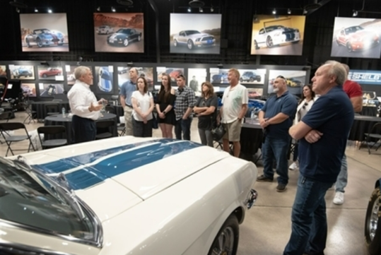 A group of people listens to a man speaking in a car museum, surrounded by classic cars and framed automotive photos on the walls.