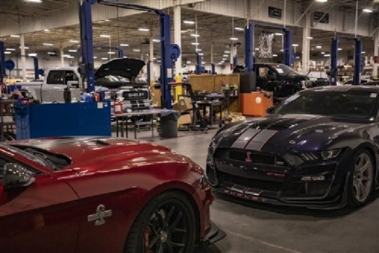 A red Shelby on the left and a blue/black on the right with a factory of car being built and worked on in the back.