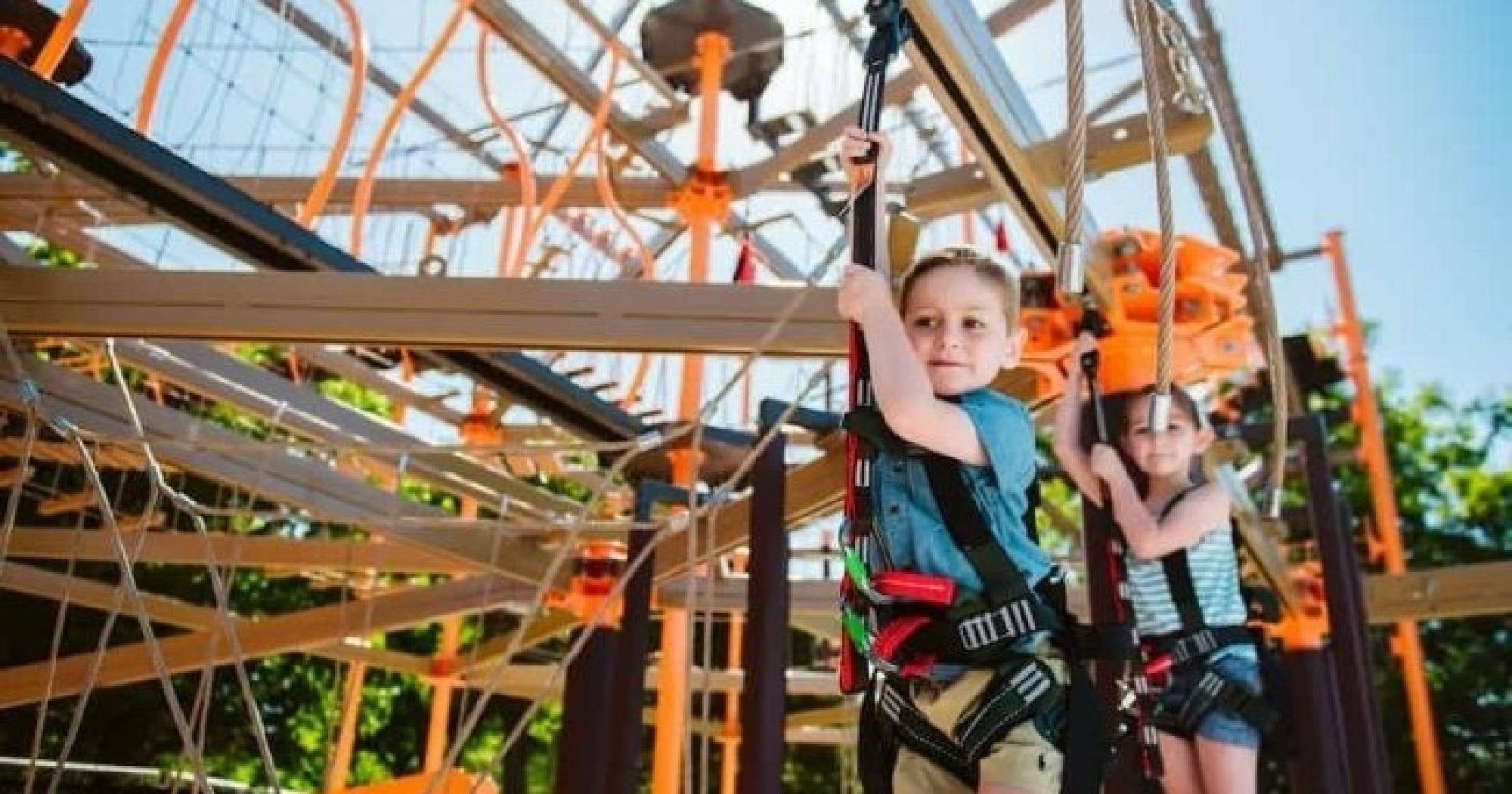 Two children wearing harnesses navigate an outdoor ropes course, holding onto safety lines with climbing structures in the background.